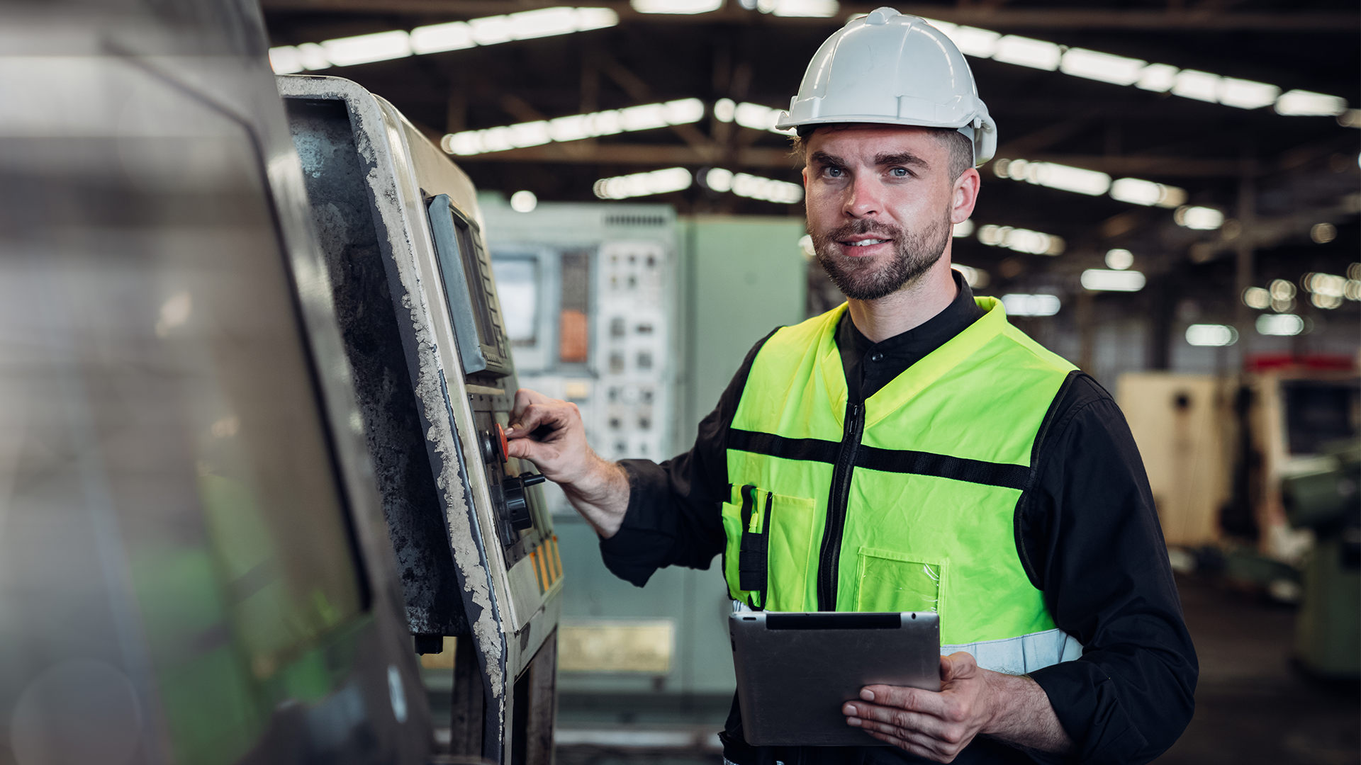 Mann in gelber Warnweste und weißen Helm bedient eine Maschine und hält ein Tablet in der Hand